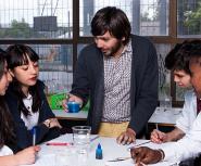 A light skinned young man with brown hair and a beard demostrates a science experiment using beekers to a diverse group of teenagers